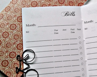 May include: A bill tracker notebook with a white interior page featuring the heading Bills, a Month line, and columns for Bill, Due Date, Amt, Paid Date, and Paid. A black metal bicycle-shaped paperclip rests on the page over a patterned cover.