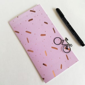 Peut inclure: Une enveloppe rectangulaire en papier violet clair ornée d'un motif dispersé de coups de pinceau et de petits points couleur cuivre métallique. Un trombone en fil de fer noir en forme de vélo est fixé à l'enveloppe, avec un stylo feutre noir.
