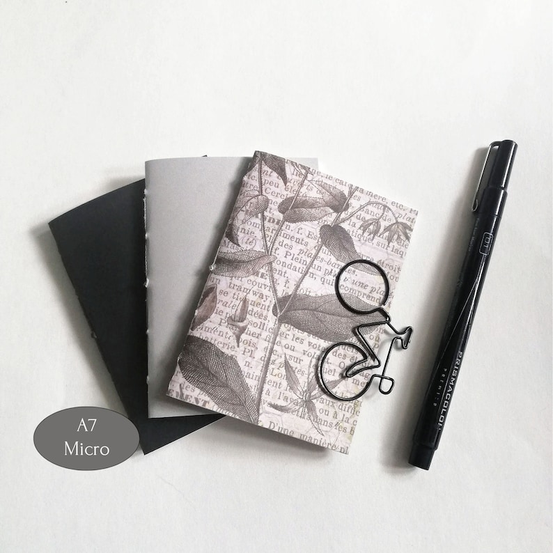 Puede incluir: Tres cuadernos peque&ntilde;os con cubiertas negras, grises y estampadas. La cubierta estampada presenta una ilustraci&oacute;n bot&aacute;nica con un clip de papel de bicicleta de metal negro. Los cuadernos est&aacute;n etiquetados como "A7 Micro".