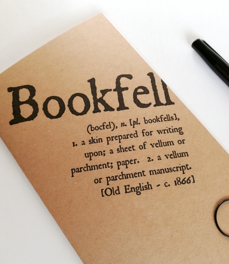 May include: A tan notebook with the word "Bookfell" printed in large, black letters. The definition of "Bookfell" is printed below in smaller, black text. A black pen is visible in the upper right corner.