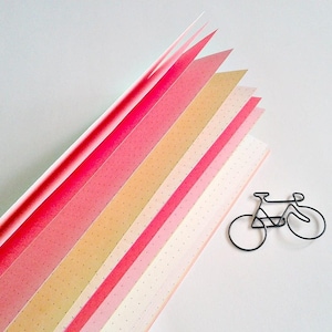 May include: A notebook with pages featuring a gradient of colours ranging from deep pink to light cream, each page marked with a subtle dot grid pattern. Beside the notebook lies a black wire paperclip shaped like a bicycle silhouette.