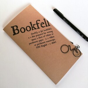 May include: A tan-colored notebook with the word "Bookfell" printed in black, along with a definition. A black pen and a bicycle-shaped paper clip are also present. The notebook is made of paper.