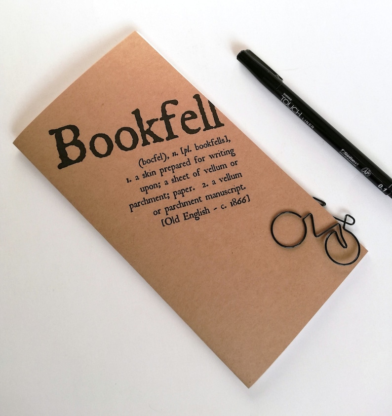 May include: A tan-colored notebook with the word "Bookfell" printed in black, along with a definition. A black pen and a bicycle-shaped paper clip are also present. The notebook is made of paper.