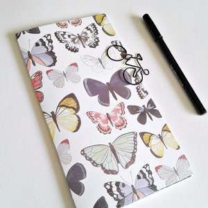 May include: A rectangular notebook with a white cover featuring a repeating pattern of butterflies in shades of purple, yellow, pink, and grey. A black metal paperclip shaped like a bicycle and a black pen are placed on top of the notebook.