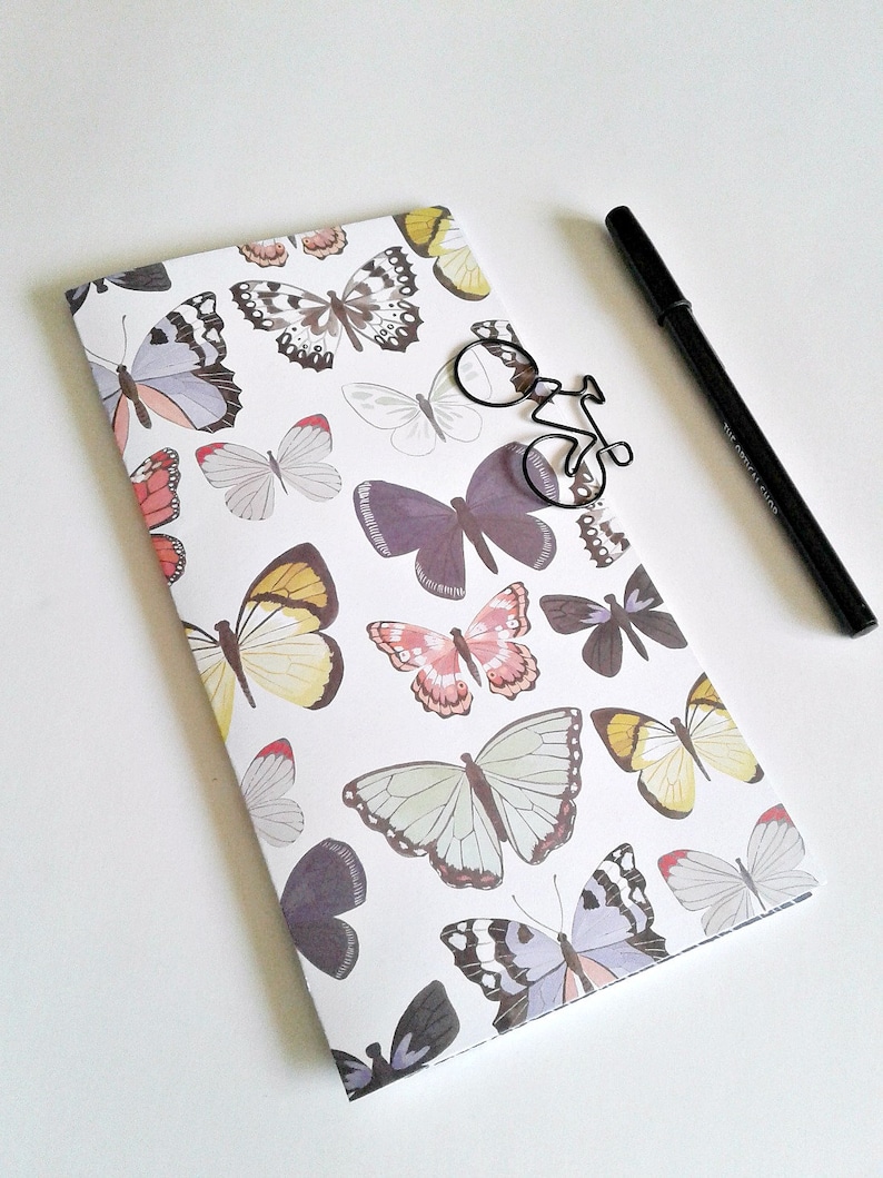 May include: A rectangular notebook with a white cover featuring a repeating pattern of butterflies in shades of purple, yellow, pink, and grey. A black metal paperclip shaped like a bicycle and a black pen are placed on top of the notebook.
