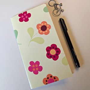 May include: A rectangular notebook with a light green cover featuring a repeating pattern of pink and orange flowers with dark centers, accompanied by a black felt-tip pen and a small black wire paperclip shaped like a bicycle.