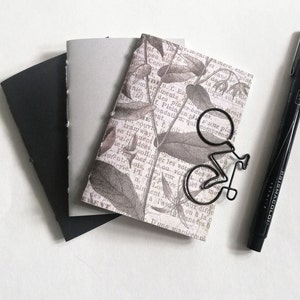 Puede incluir: Tres cuadernos peque&ntilde;os con tapas negras, grises y estampadas. La tapa estampada presenta un dise&ntilde;o floral con un marcap&aacute;ginas de metal negro en forma de bicicleta. Tambi&eacute;n se ve un rotulador negro en la imagen.