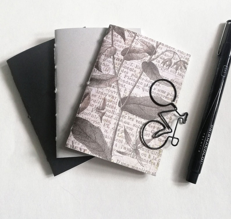 Puede incluir: Tres cuadernos peque&ntilde;os con tapas negras, grises y estampadas. La tapa estampada presenta un dise&ntilde;o floral con un marcap&aacute;ginas de metal negro en forma de bicicleta. Tambi&eacute;n se ve un rotulador negro en la imagen.