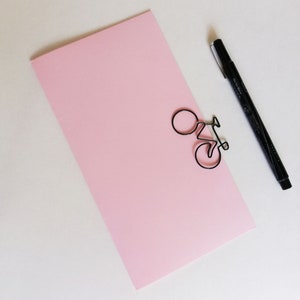 Peut inclure: Une carte rectangulaire en papier rose clair avec un trombone en fil de fer noir en forme de vélo. Un stylo marqueur noir à pointe fine avec son capuchon est placé en diagonale à côté de la carte. Les objets sont sur une surface neutre.