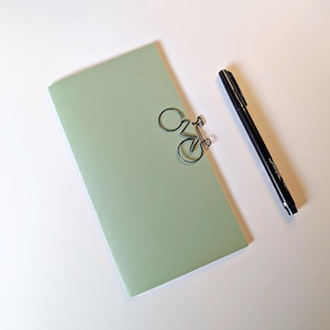 Peut inclure: Un carnet rectangulaire vert sauge clair avec un trombone en fil de fer noir en forme de vélo fixé sur le bord supérieur, accompagné d'un stylo-feutre noir fin positionné en diagonale à droite du carnet sur une surface unie.