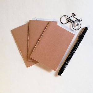 Puede incluir: Tres cuadernos marrones con costuras a lo largo del lomo, un marcador negro y una silueta de bicicleta de alambre negro.