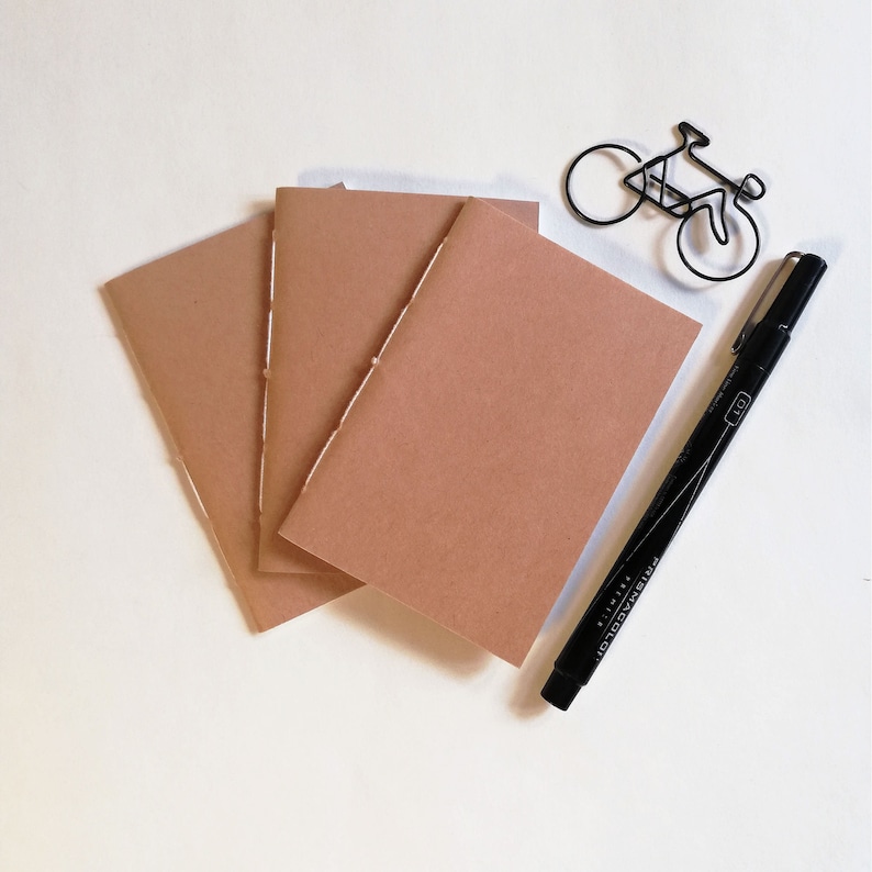 Puede incluir: Tres cuadernos marrones con costuras a lo largo del lomo, un marcador negro y una silueta de bicicleta de alambre negro.