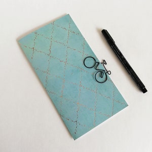Peut inclure: Un carnet rectangulaire bleu clair avec un motif de losanges pointillés couleur cuivre, un trombone en fil de fer noir en forme de vélo fixé sur la couverture et un feutre noir placé en diagonale à côté du carnet sur une surface blanche.