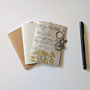 May include: Two small notebooks and a black pen. One notebook has a plain brown cover, while the other features a vintage-style paper cover with cursive script text, yellow flower illustrations, and a black wire bicycle-shaped paper clip.