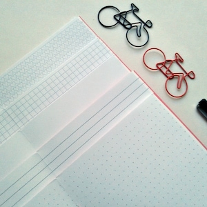 May include: A notebook with various page styles including heart patterns, grid lines, horizontal lines, and dot grids, accompanied by one black bicycle-shaped paper clip, one red bicycle-shaped paper clip, and a black pen tip.