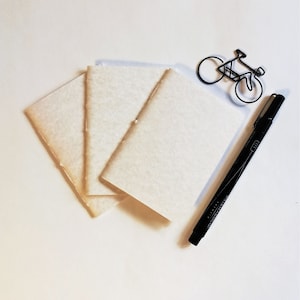 Puede incluir: Una pila de tres cuadernos de papel blanco crema en blanco con un bol&iacute;grafo negro y una silueta de bicicleta de alambre sobre un fondo blanco.