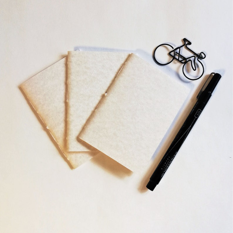 Puede incluir: Una pila de tres cuadernos de papel blanco crema en blanco con un bol&iacute;grafo negro y una silueta de bicicleta de alambre sobre un fondo blanco.