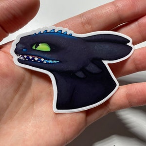 May include: A black and blue cartoon dragon sticker with a green eye. The dragon is facing to the right and has a toothy grin.