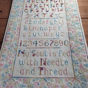 May include: A white cross-stitch sampler with a colorful alphabet, numbers, and the phrase "My Soul is fed with Needle and Thread".