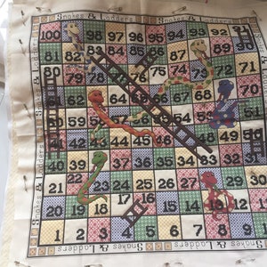 May include: A fabric game board for the game Snakes and Ladders. The board is beige with colourful squares and features images of snakes and ladders. The numbers 1 through 100 are printed on the squares. The text "Snakes & Ladders" is printed on the top and bottom of the board.