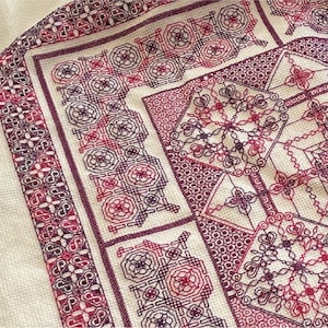 Puede incluir: Primer plano de una tela blanca con un diseño de bordado detallado en tonos rojos y morados. El diseño presenta intrincados patrones geométricos y motivos florales.