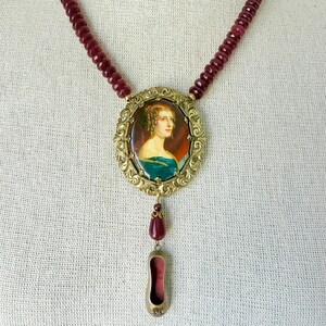 May include: A vintage necklace featuring a portrait pendant. The pendant is framed in gold-toned metal and hangs from a strand of burgundy beads. A teardrop-shaped charm and a shoe-shaped charm dangle below the portrait.