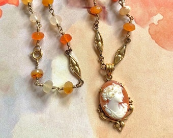 May include: A gold chain necklace with a cameo pendant featuring a woman's profile. The chain is adorned with orange and white beads.