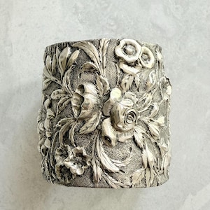 May include: A silver-toned cuff bracelet with an intricate floral design. The detailed pattern features roses, leaves, and other flowers in a raised relief. The cuff has a textured background, giving it a vintage or antique appearance.
