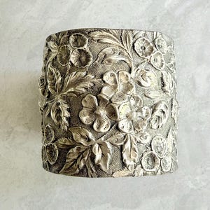 May include: Silver-toned cuff bracelet with an embossed floral design. The cuff features detailed relief work of flowers and leaves, creating a textured surface. The bracelet has a wide band and a vintage aesthetic.