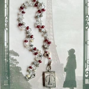 May include: A necklace with a silver pendant featuring the Eiffel Tower, strung with a mix of red, gray, and white beads. The necklace is displayed against a backdrop of the Eiffel Tower and a vintage-style image.