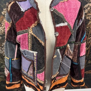 Könnte beinhalten: Eine langärmelige Jacke aus einem Patchwork verschiedener Stoffe wie Samt, Tweed und Brokat in den Farben Pink, Lila, Braun, Schwarz und Orange. Die Jacke verfügt über eine Knopfleiste auf der Vorderseite mit vier runden Knöpfen.