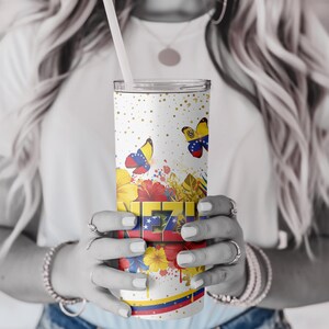 May include: A white tumbler with a straw, featuring a design of butterflies, flowers, and gold speckles. The tumbler has the word "VE" in a colorful, blocky font, with the Venezuelan flag colors. A decorative sleeve is wrapped around the tumbler.