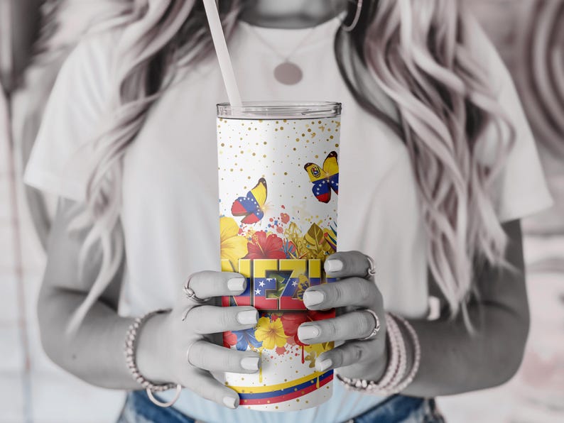 May include: A white tumbler with a straw, featuring a design of butterflies, flowers, and gold speckles. The tumbler has the word "VE" in a colorful, blocky font, with the Venezuelan flag colors. A decorative sleeve is wrapped around the tumbler.