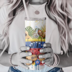 May include: A white tumbler with a straw, featuring a colorful design with Venezuelan flag elements, musical instruments, and tropical flowers. The design includes a guitar, maracas, and a coffee mug. The tumbler is held by a person.