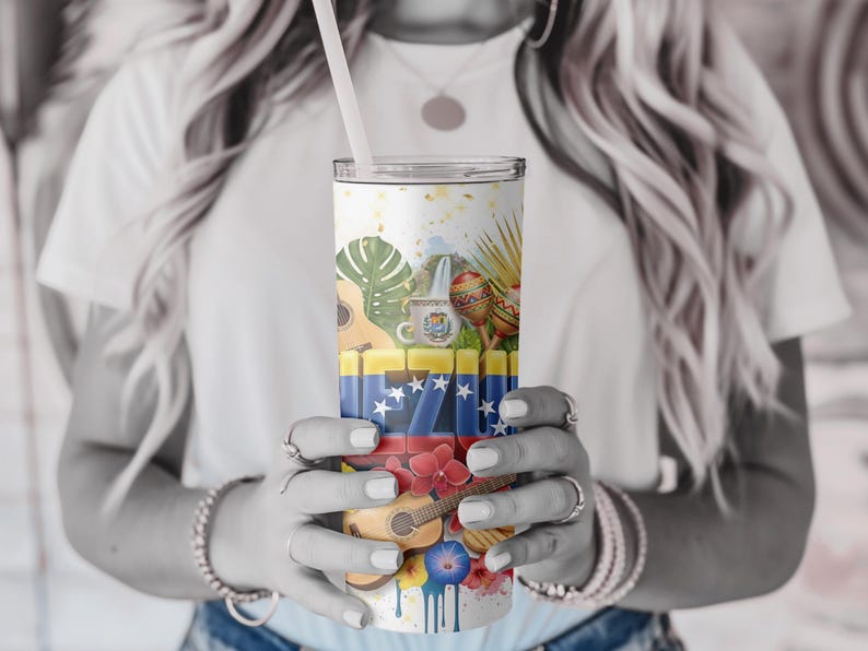 May include: A white tumbler with a straw, featuring a colorful design with Venezuelan flag elements, musical instruments, and tropical flowers. The design includes a guitar, maracas, and a coffee mug. The tumbler is held by a person.