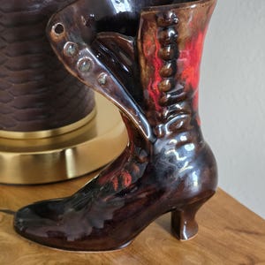 May include: A ceramic boot-shaped vase with a glossy finish in shades of dark brown and deep red. The design features a folded-over cuff, a row of molded buttons along the front, and a small heel. The interior is hollow, forming a functional vessel.