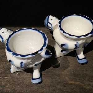 May include: Two white ceramic egg cups featuring a blue hand-painted scalloped rim and blue accents. Each cup is shaped like a small figure with a head, arms, and legs, with the egg holder positioned in the center of the body.