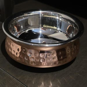 May include: A copper-colored bowl with a hammered texture on the exterior and a shiny silver interior. The bowl has a wide, rounded shape and a polished silver rim. The bowl is likely used for serving food.