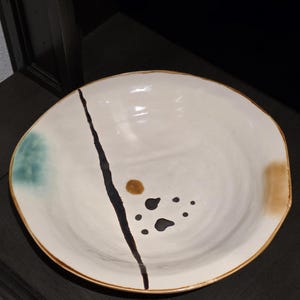 May include: A large, off-white ceramic bowl with a brown rim and abstract black and brown painted details. A dark brown line bisects the bowl, with a teal and brown accent on the sides.