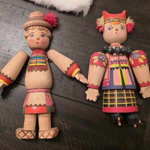 May include: Two wooden dolls with painted features and articulated limbs. The left doll wears a hat with a pink pom-pom and a patterned tunic. The right doll features a headpiece with pink pom-poms, a beaded necklace, and a plaid skirt design.