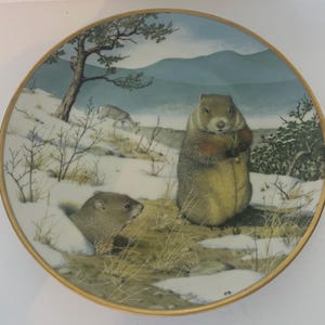 May include: A decorative ceramic plate with a winter scene featuring two groundhogs, one standing and one peeking out of a burrow, in a snowy landscape with a pine tree and a deer in the background.
