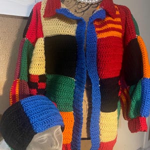 May include: A colorful, patchwork crochet jacket and matching beanie. The jacket features a collared neckline and a blue, open front. The beanie is color-blocked in black, blue, and cream. The jacket and beanie are displayed on a mannequin.
