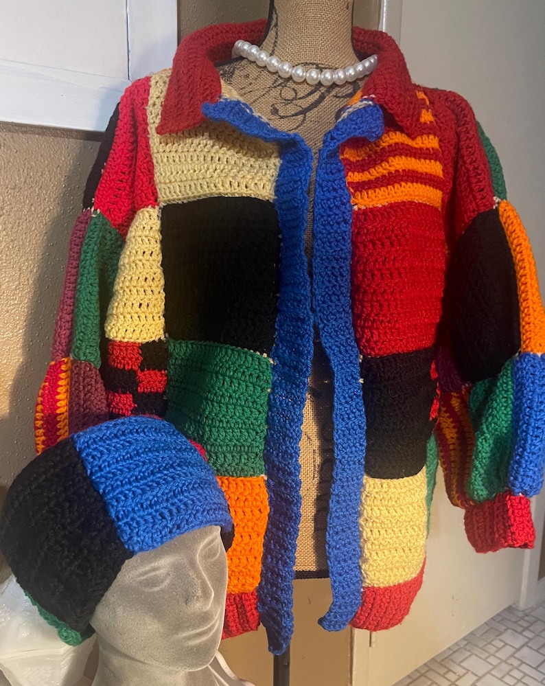 K&ouml;nnte beinhalten: Eine farbenfrohe Patchwork-H&auml;keljacke und eine passende M&uuml;tze. Die Jacke hat einen Kragen und eine blaue, offene Vorderseite. Die M&uuml;tze ist in Schwarz, Blau und Creme gehalten. Jacke und M&uuml;tze werden auf einer Schaufensterpuppe pr&auml;sentiert.