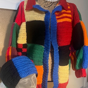 May include: A colorful, patchwork crochet jacket and matching beanie. The jacket features a collared neckline and a blue, open front. The beanie is color-blocked in black, blue, and cream. The jacket and beanie are displayed on a mannequin.