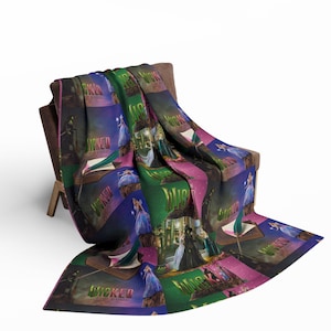 Peut inclure: Une couverture colorée drapée sur une chaise en bois. La couverture présente un motif répété avec le mot "Wicked" en vert et des images de personnages d'une production de théâtre musical. L'arrière-plan comprend des nuances de violet, de rose et de vert.