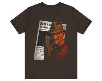 Könnte beinhalten: Dunkelbraunes T-Shirt mit einer Grafik von Freddy Krueger aus der Filmreihe Nightmare on Elm Street. Die Grafik enthält einen Zeitungsausschnitt mit der Überschrift "ELM STREET TERROR STRIKES".