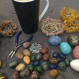 May include: A collection of colorful stones, turquoise, and agate, arranged on a gray surface with a blue mug, a pair of pliers, and wire sculptures in the shape of nests.