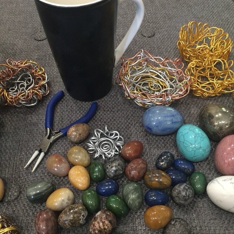 May include: A collection of colorful stones, turquoise, and agate, arranged on a gray surface with a blue mug, a pair of pliers, and wire sculptures in the shape of nests.