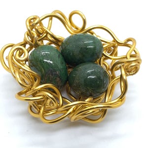May include: A gold wire nest with three green gemstone eggs. The nest is made of twisted wire and the eggs are smooth and shiny.
