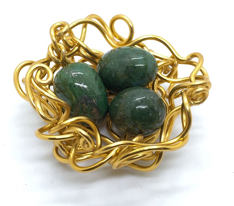 May include: A gold wire nest with three green gemstone eggs. The nest is made of twisted wire and the eggs are smooth and shiny.